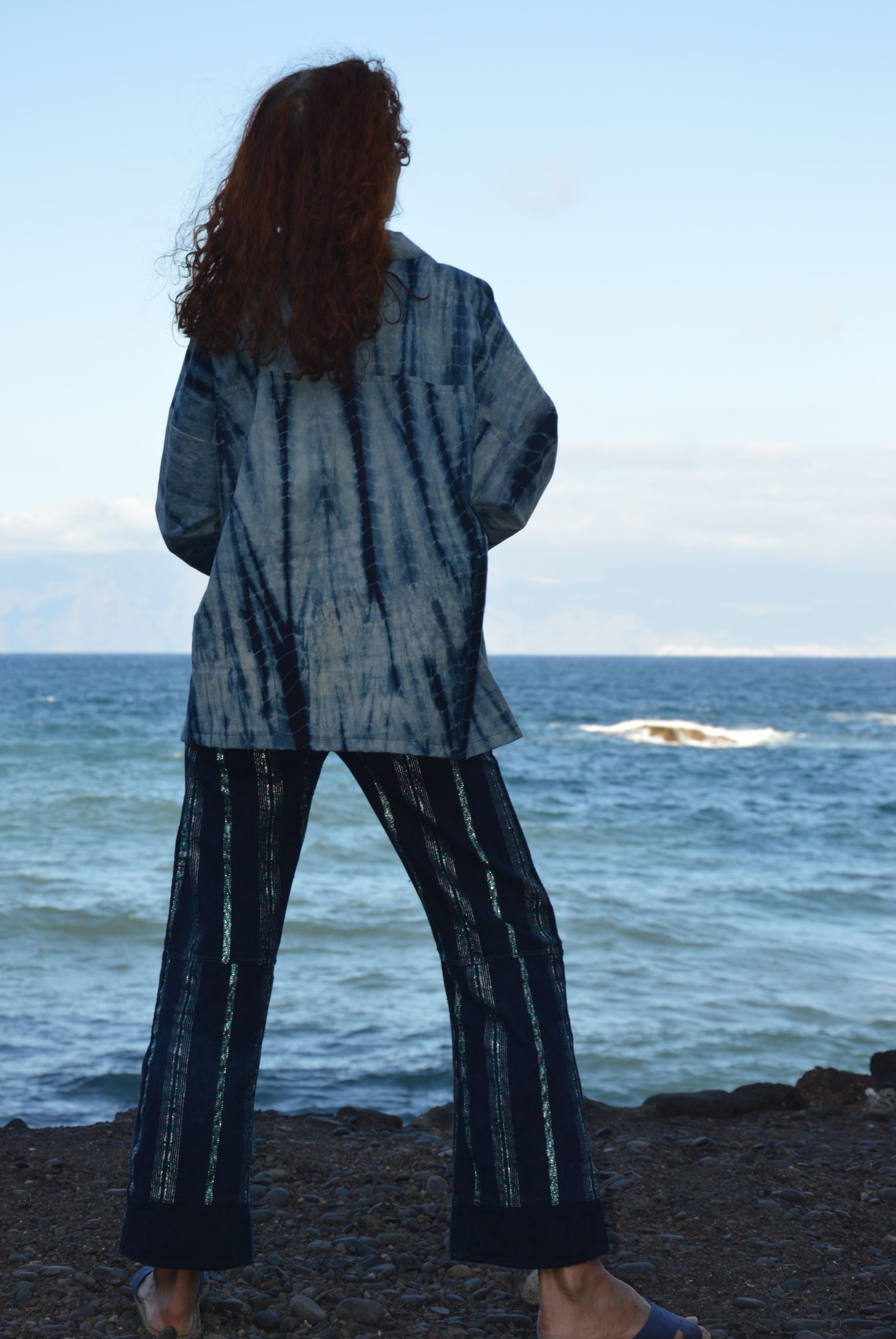 Persona de pie en una costa rocosa frente al océano, vestida con una chaqueta azul y pantalones oscuros.