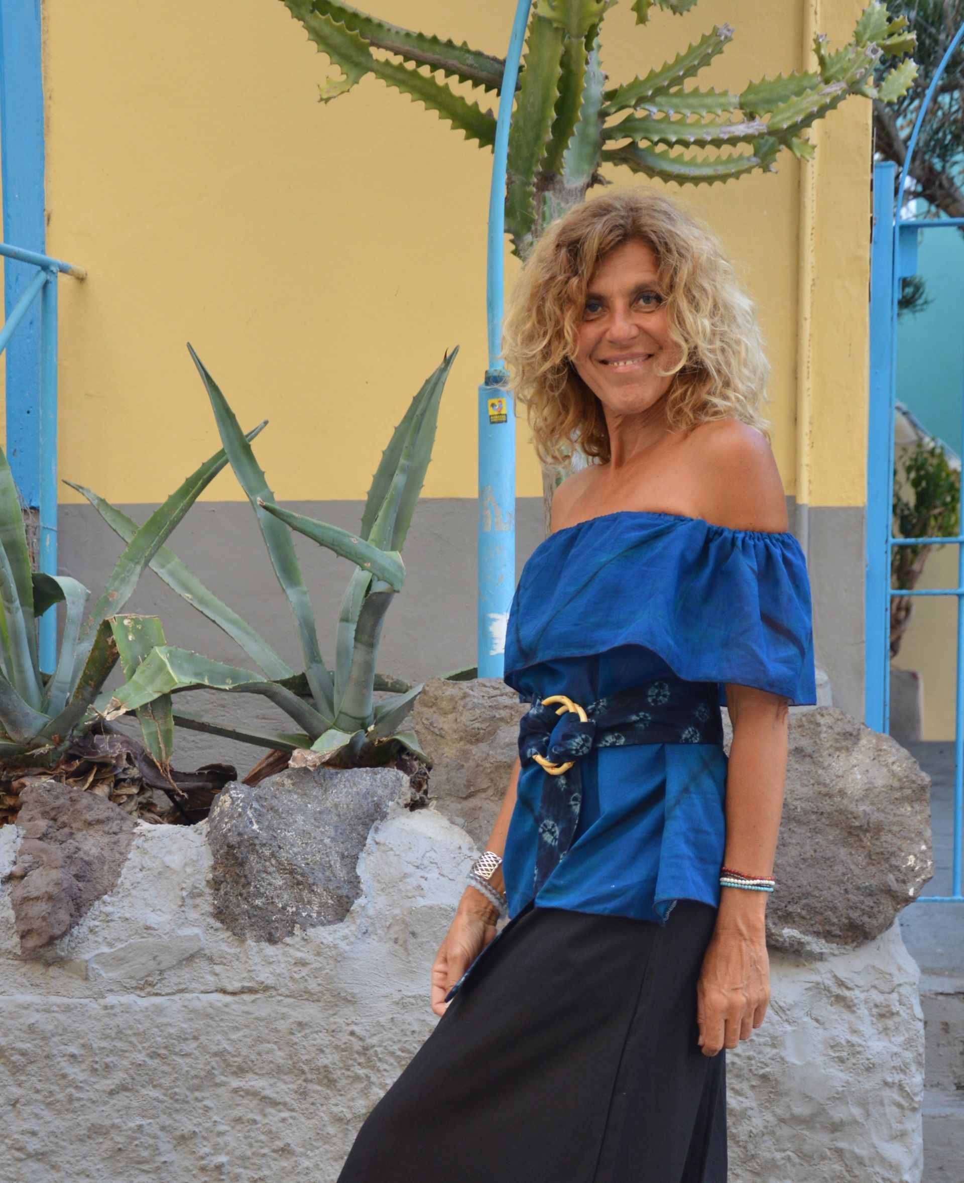 Mujer con top azul sin hombros y falda negra de pie junto a una pared amarilla y plantas de agave.
