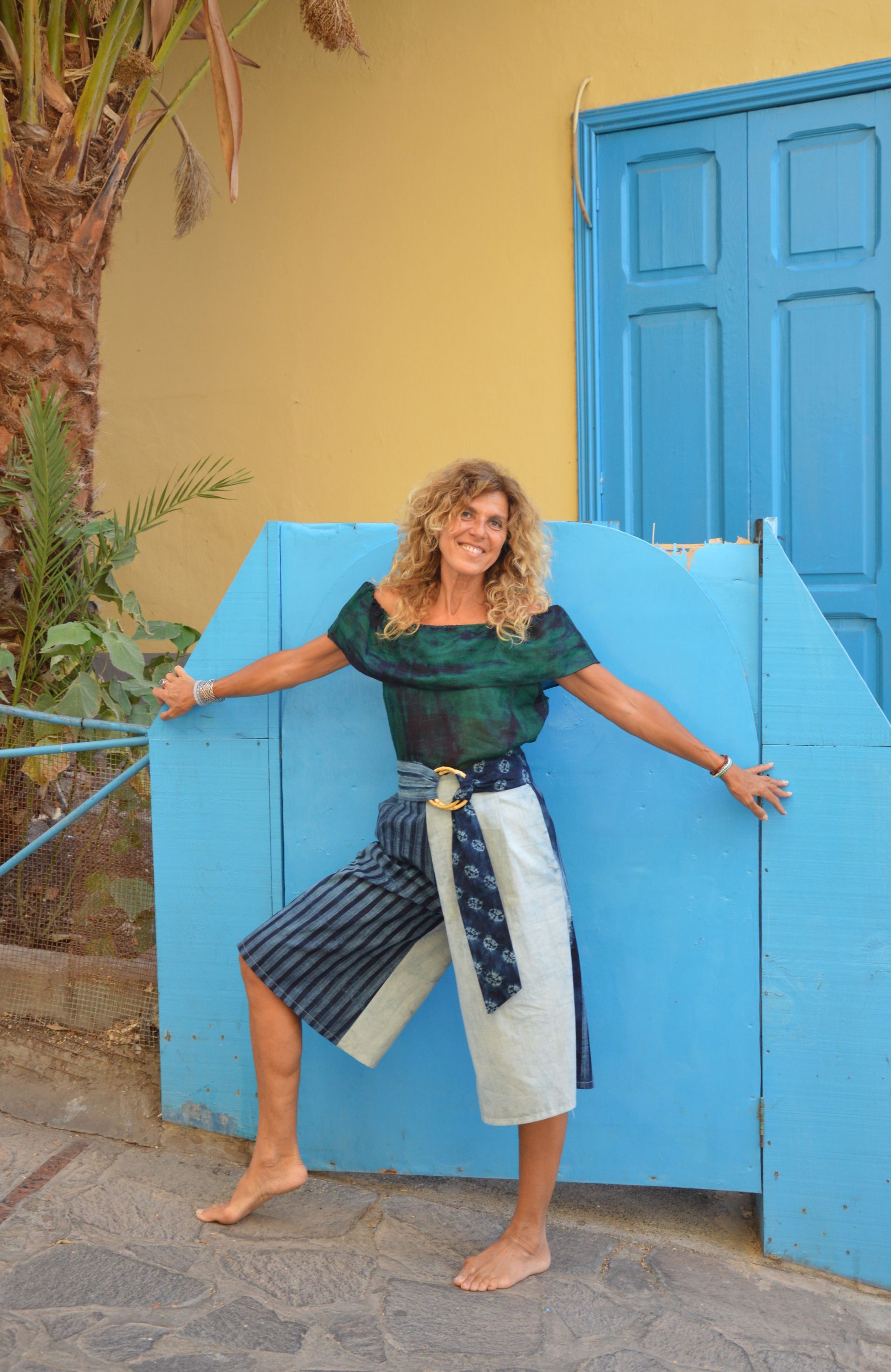 Mujer sonriendo y posando con los brazos extendidos junto a una puerta azul brillante y una pared amarilla.