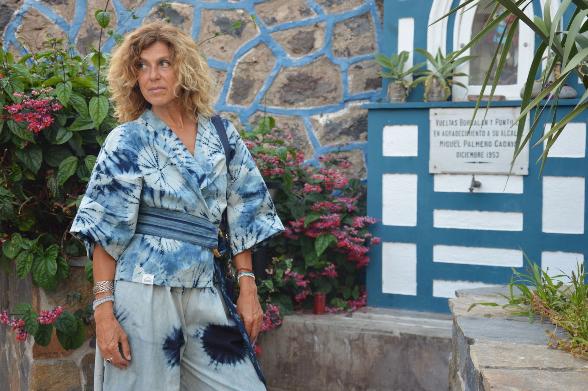Mujer con un atuendo azul teñido con la técnica tie-dye de pie junto a una pared azul y blanca y flores al aire libre.