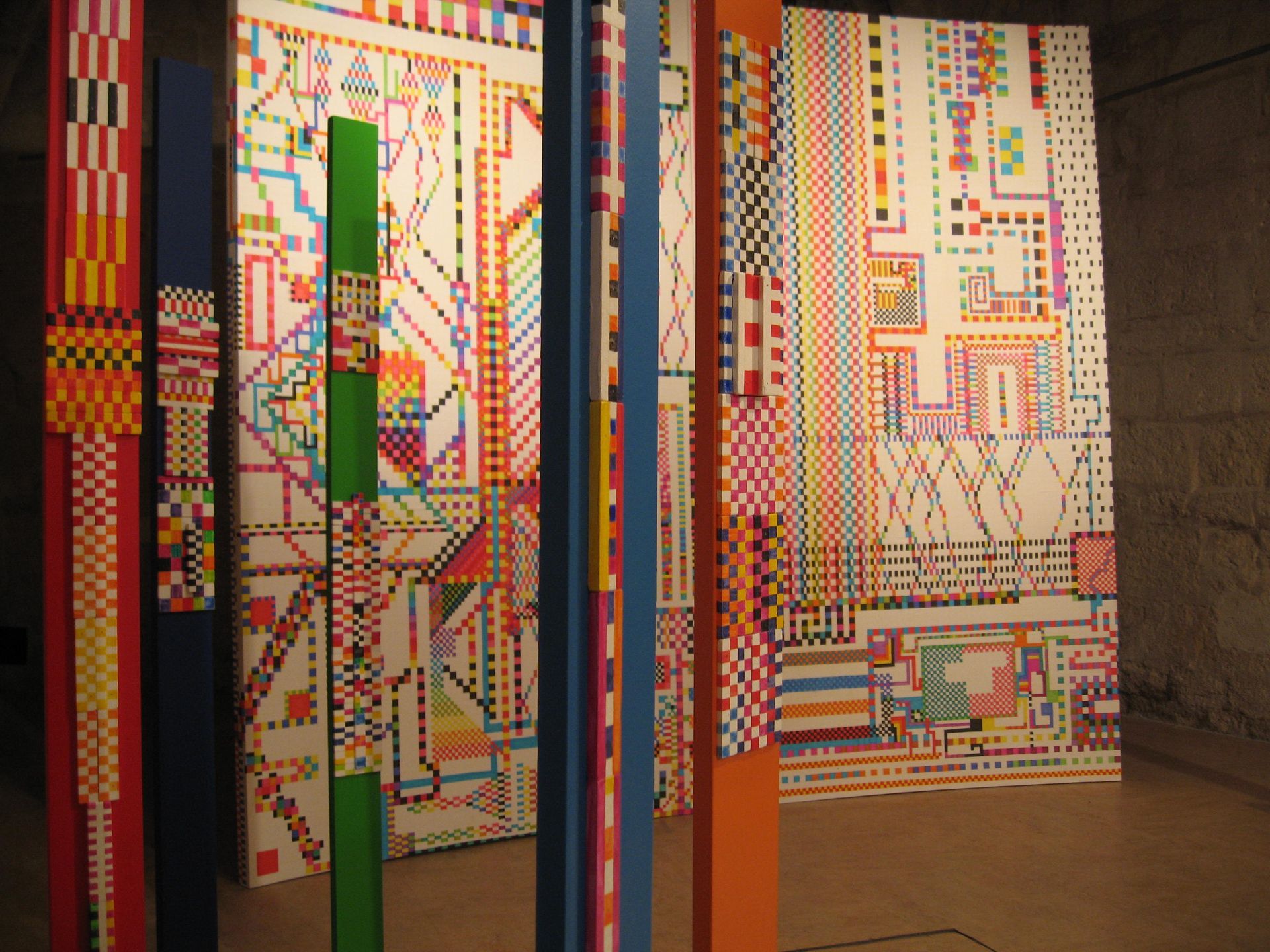 Cinco esculturas altas y verticales, cubiertas de coloridos motivos geométricos, se alzan en una habitación con paredes de textura rugosa similar a la piedra.