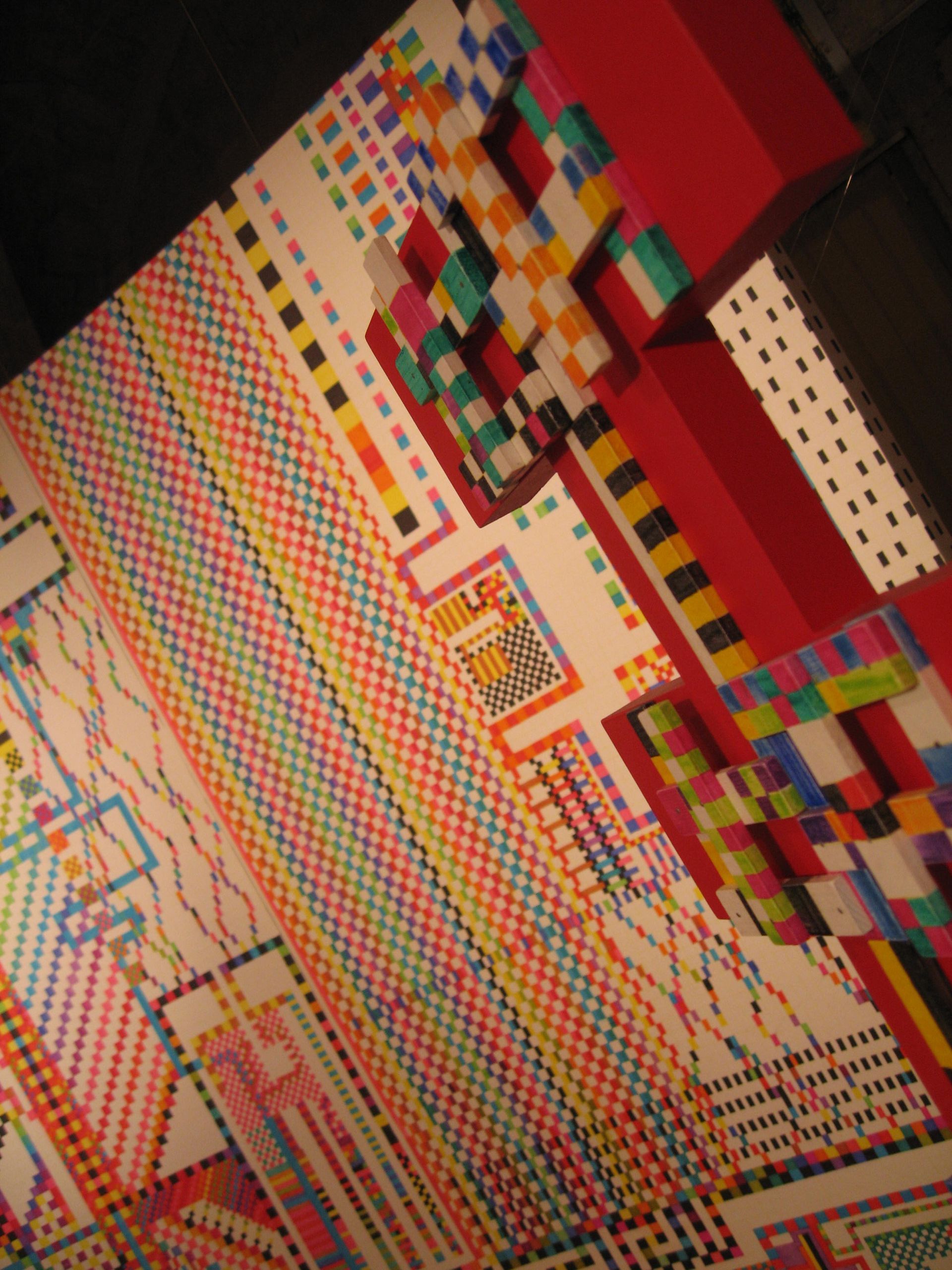 Detrás de dos barras verticales de color azul oscuro y verde, se encuentra un mural con coloridos patrones geométricos estilo píxel.