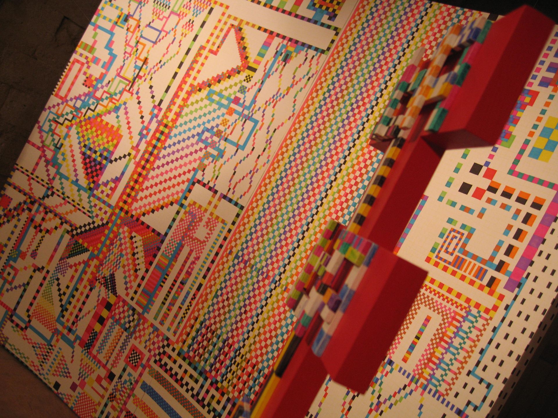 Un mural abstracto y colorido con patrones geométricos repetitivos, bloques pixelados y líneas sobre un fondo blanco.