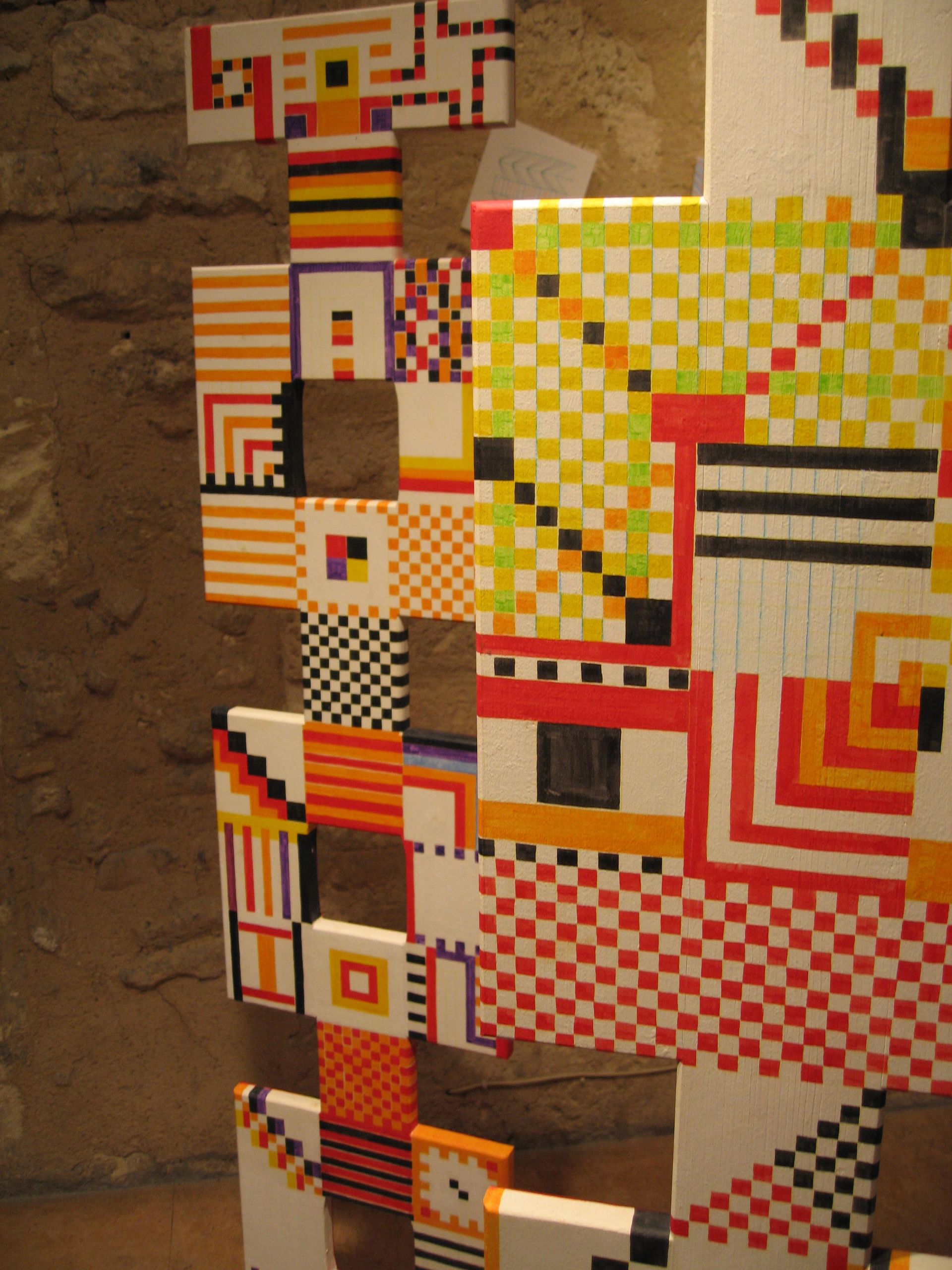 Escultura geométrica abstracta con variados patrones coloridos en forma de cuadrícula sobre paneles blancos, colocada sobre una pared de hormigón.