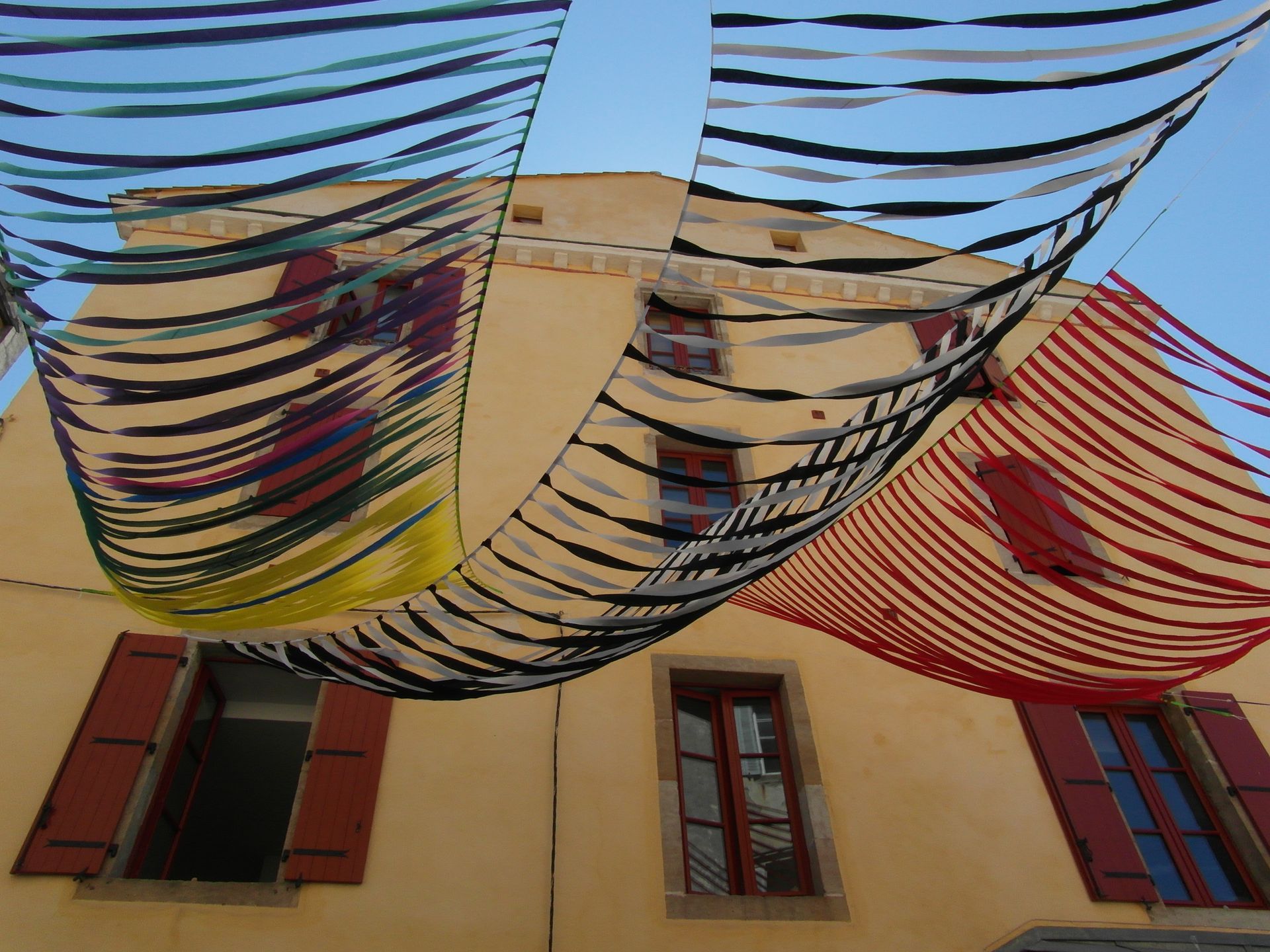 Cintas de colores cuelgan en hileras entre un edificio amarillo y el cielo, creando un dosel festivo.