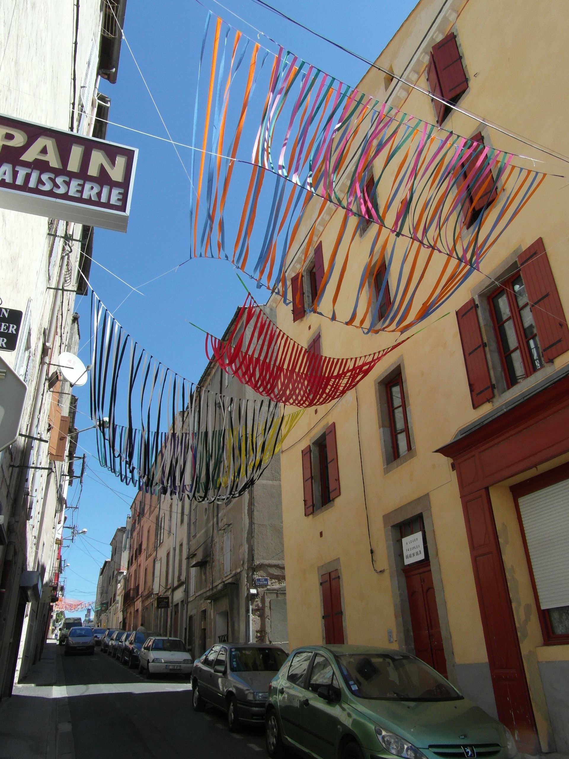 Tiras decorativas de tela azul, verde y roja cuelgan sobre un edificio amarillo con contraventanas de color rojo oscuro.