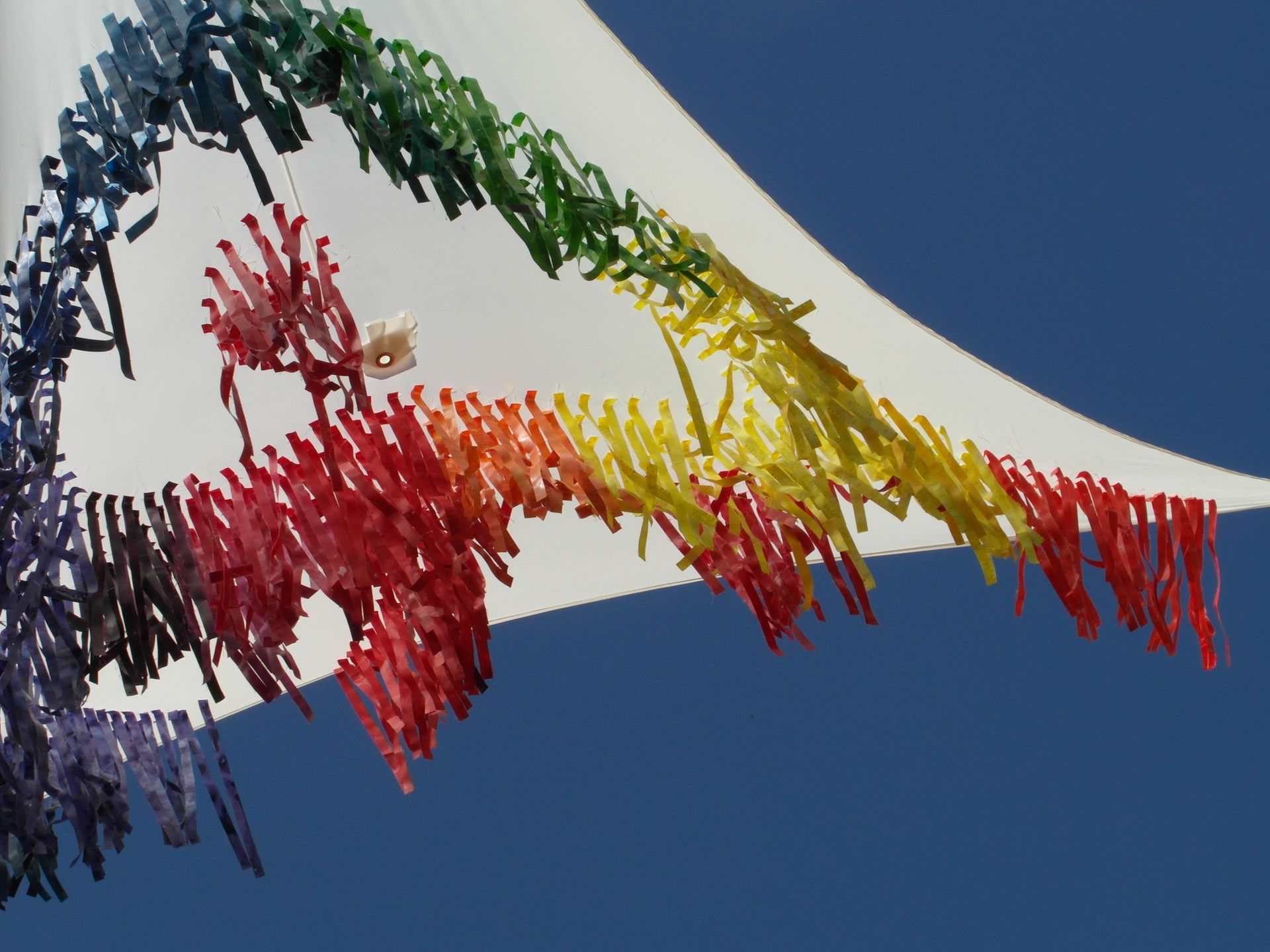 Una colorida escultura abstracta hecha de cintas o tiras colgantes, que caen en cascada creando un degradado de arcoíris sobre un cielo azul.