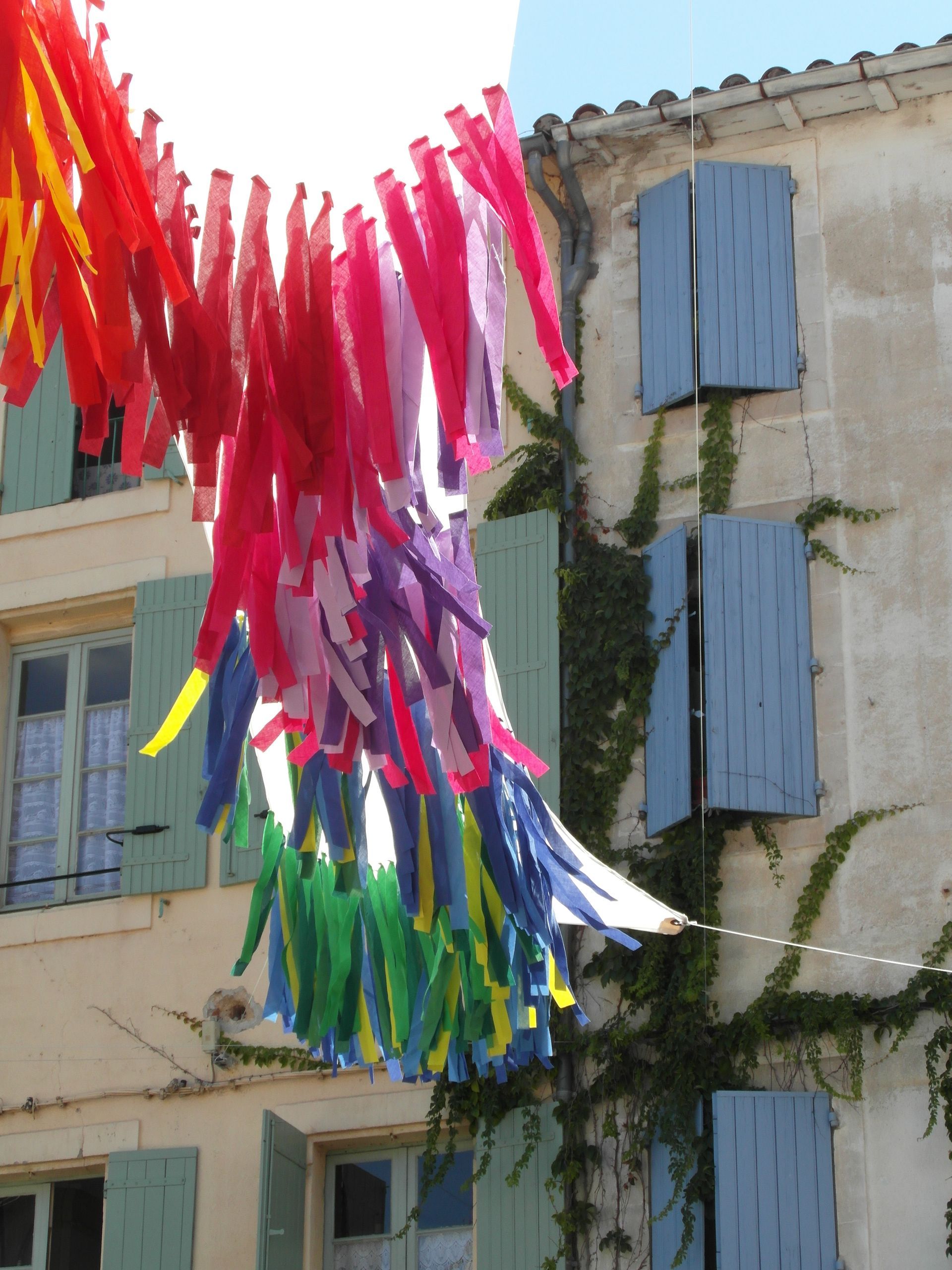 Tiras de tela de colores cuelgan como banderines entre viejos edificios de piedra con contraventanas azules y hiedra trepadora.