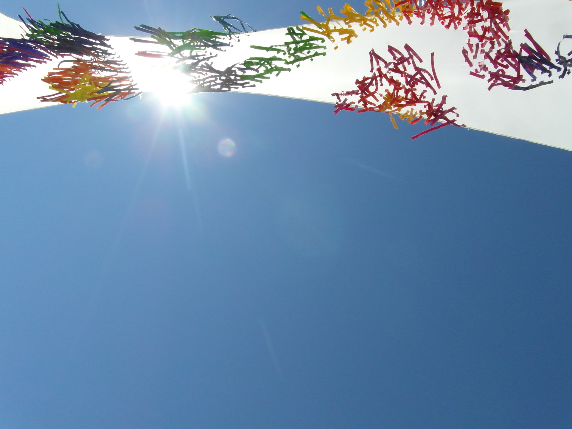 Un sol radiante brilla tras un arco blanco decorado con coloridas guirnaldas colgantes, contra un cielo azul despejado.