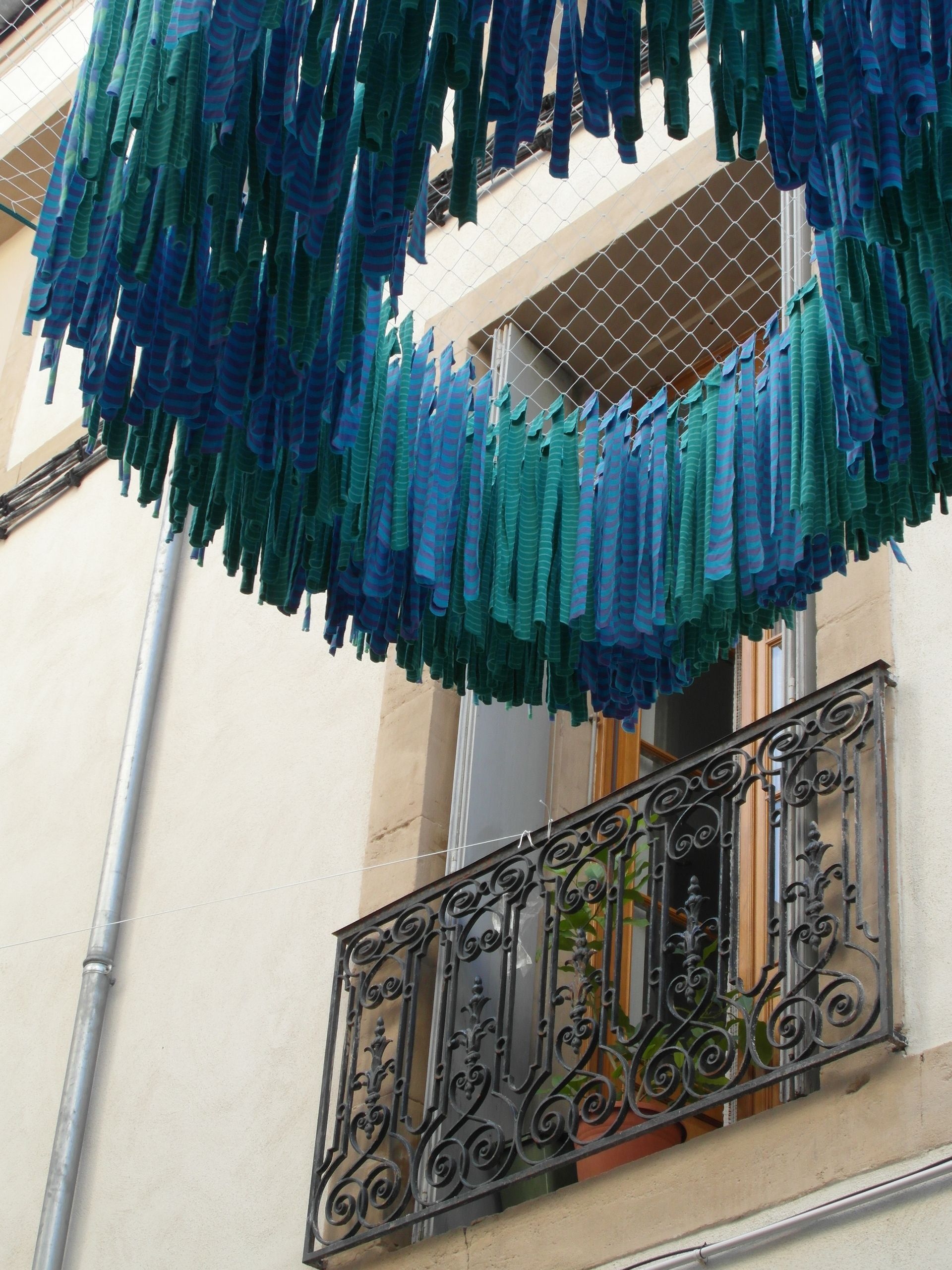 Una instalación circular de tiras de tela azules y turquesas que cuelgan sobre un balcón con una barandilla de hierro ornamentada.