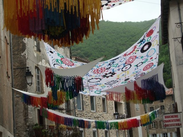 Doseles de tela blanca estampados con diseños coloridos y flecos de colores del arcoíris cuelgan entre edificios históricos de piedra.