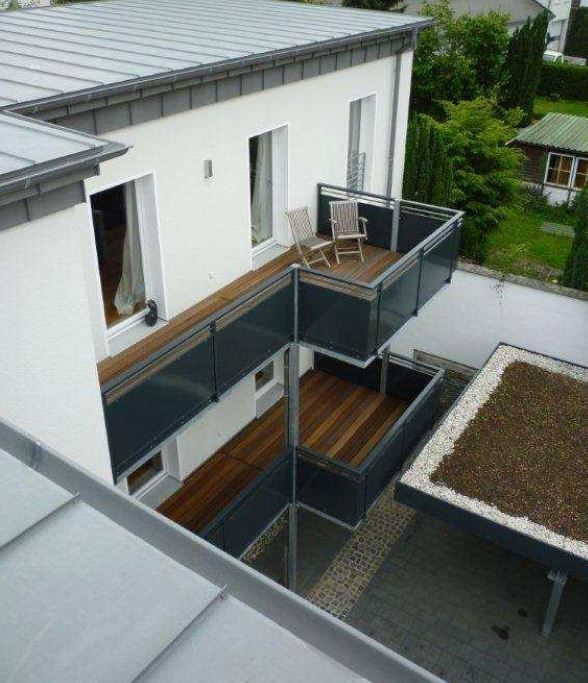 Modernes weißes Haus mit grauen Metallverzierungen, einem Holzbalkon mit Stuhl und einem Gründach.