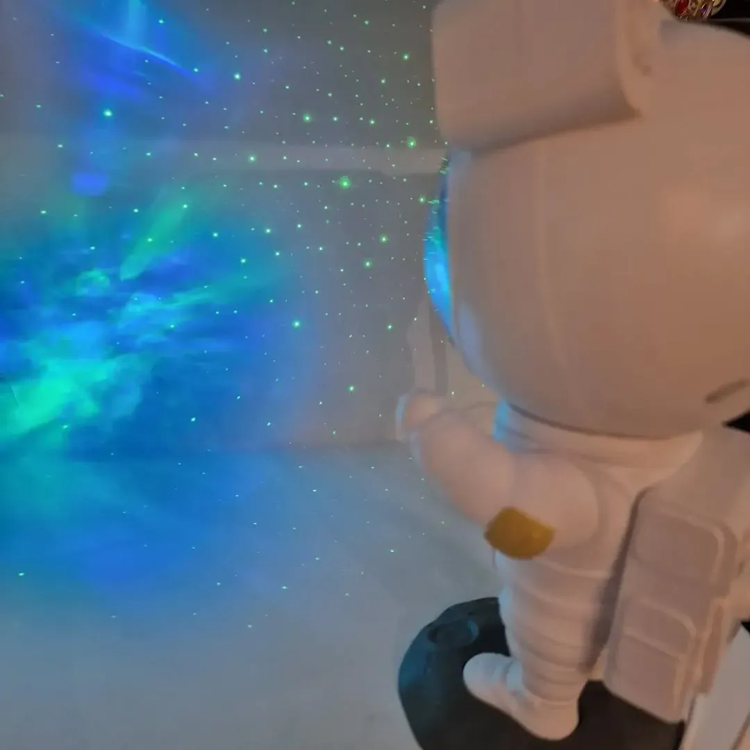 Un proyector con forma de astronauta ilumina una pared con nubes de nebulosas azules y verdes en espiral