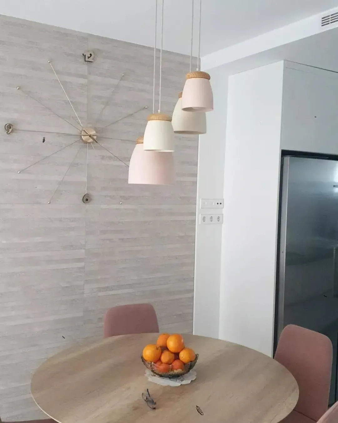 Una mesa de comedor de madera con un cuenco de naranjas, tres lámparas colgantes y un reloj decorativo