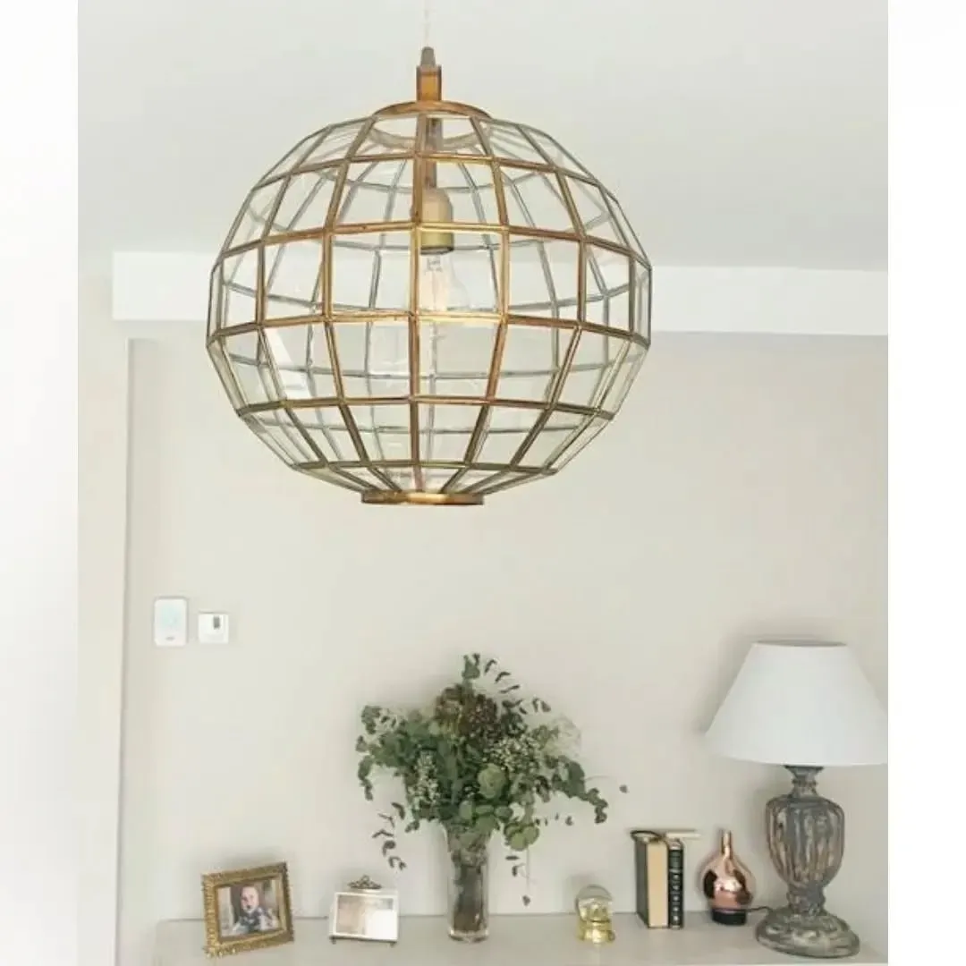 Una lámpara colgante esférica de cristal con marco dorado cuelga sobre una mesa consola con un jarrón con plantas