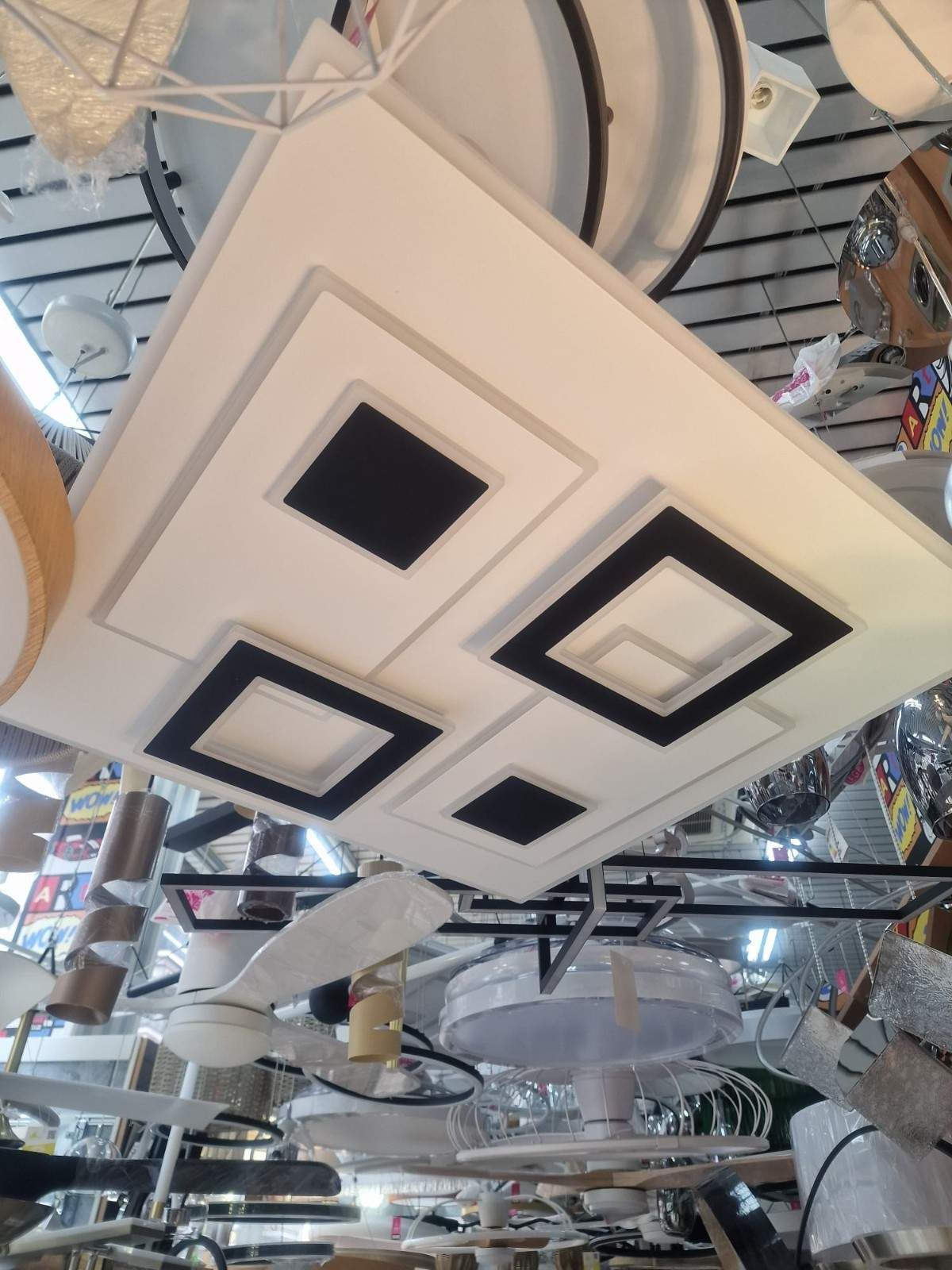 Una lámpara de techo LED cuadrada, moderna, blanca y negra, cuelga en una sala de exposición repleta de otras opciones