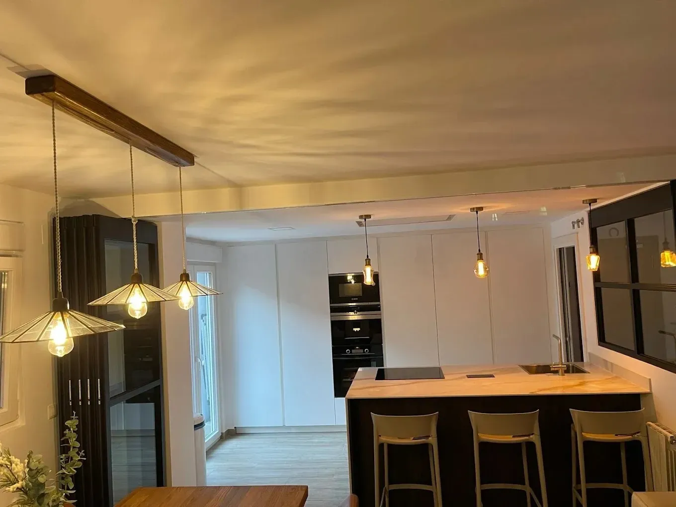 Cocina moderna con una isla de color claro, tres taburetes de bar, armarios blancos e iluminación