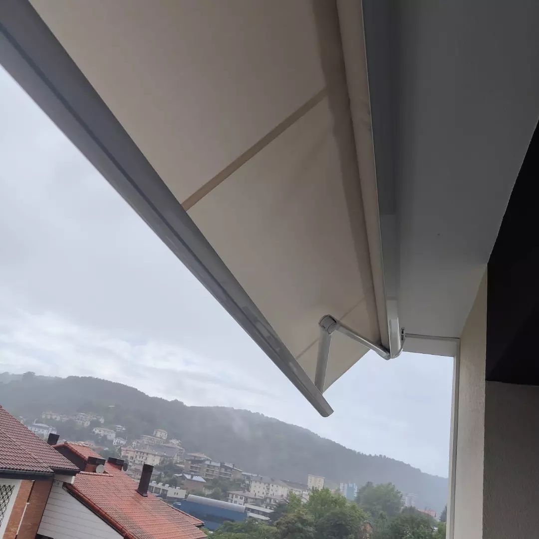 Un toldo retráctil de color claro se extiende sobre un balcón. Al fondo se ve un pueblo en la ladera bajo un cielo nublado.