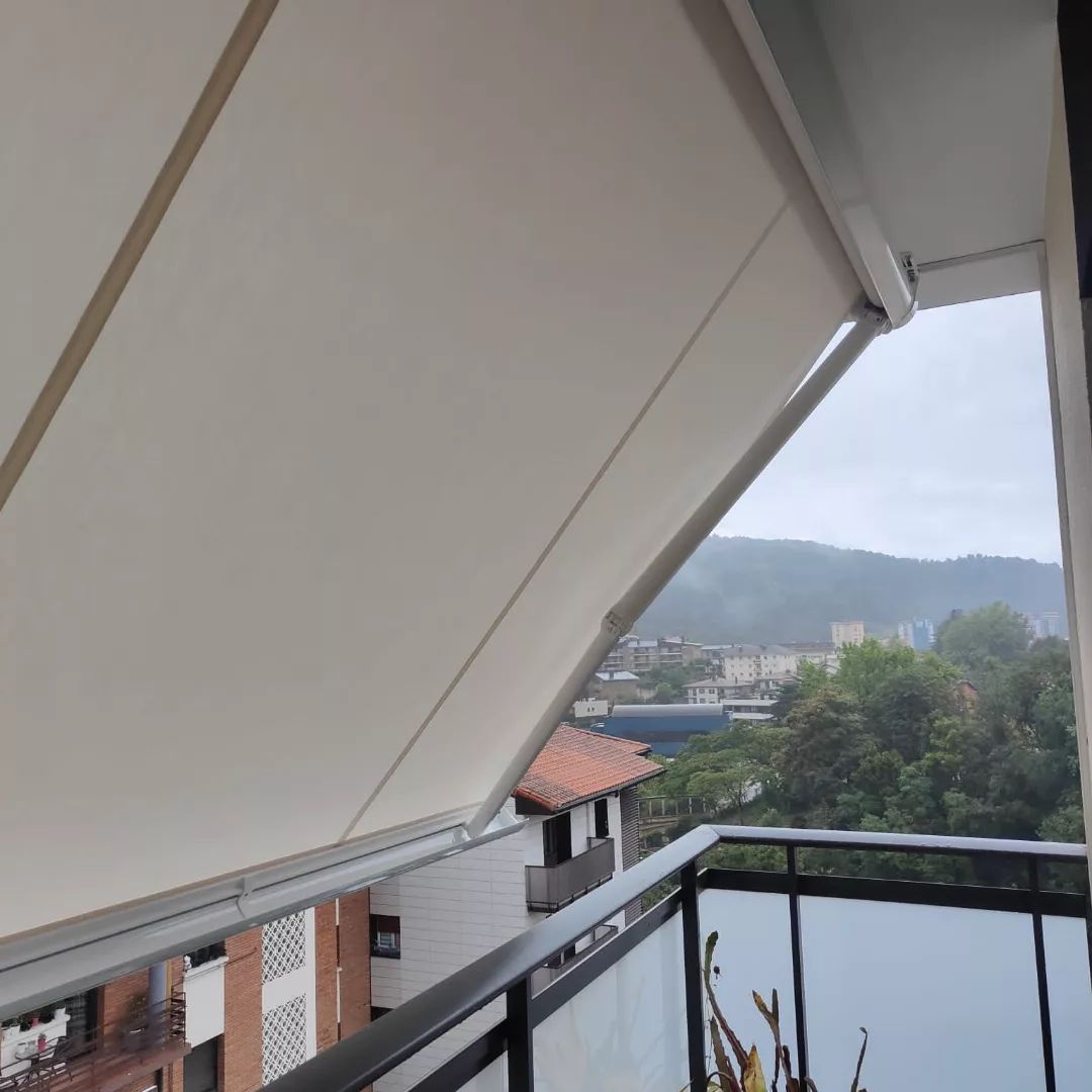 Un toldo blanco parcialmente desplegado en un balcón, con vistas a una ciudad y montañas en un día nublado.