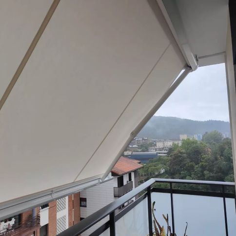 Un toldo blanco parcialmente extendido sobre un balcón, con vistas a una ciudad y colinas verdes bajo un cielo nublado.