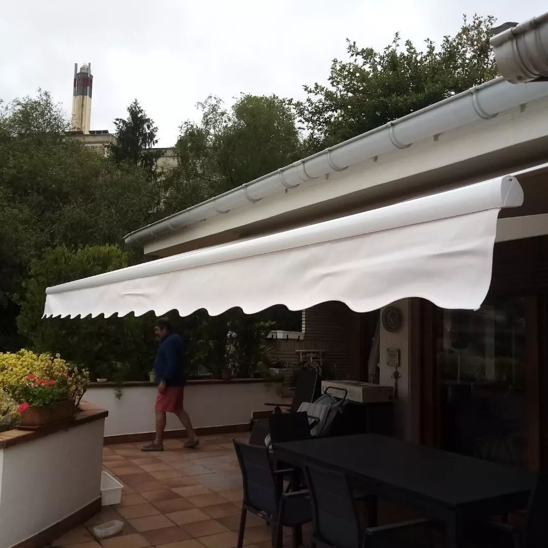 Un toldo blanco da sombra a un patio con mesa y sillas; una persona camina cerca. Al fondo se ven árboles y un edificio.