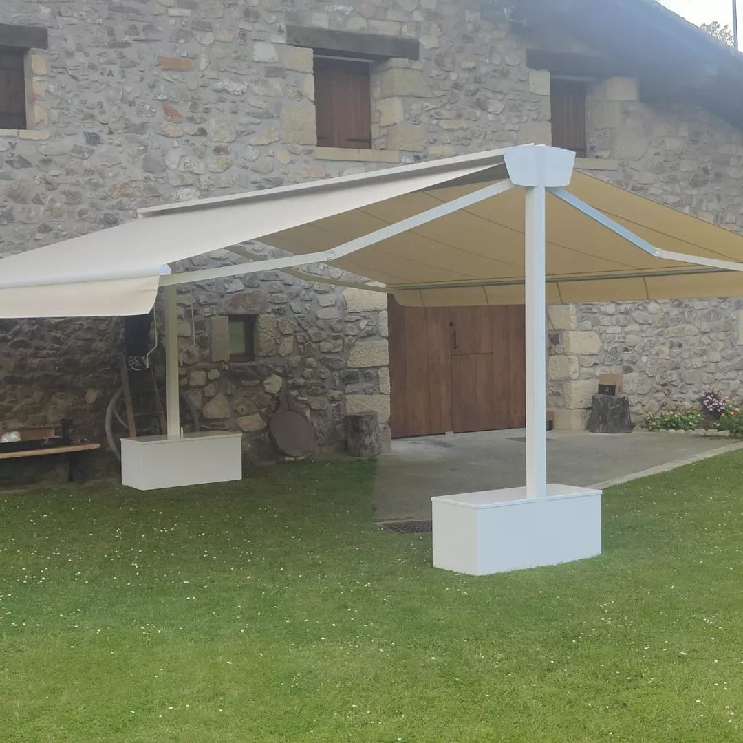 Una gran sombrilla de color beige con soporte blanco se encuentra sobre un césped frente a un edificio de piedra con puertas de madera.