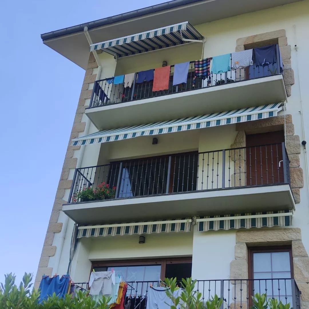 Fachada de edificio con tres balcones, toldos de rayas verdes y blancas, y ropa tendida. Cielo azul arriba.