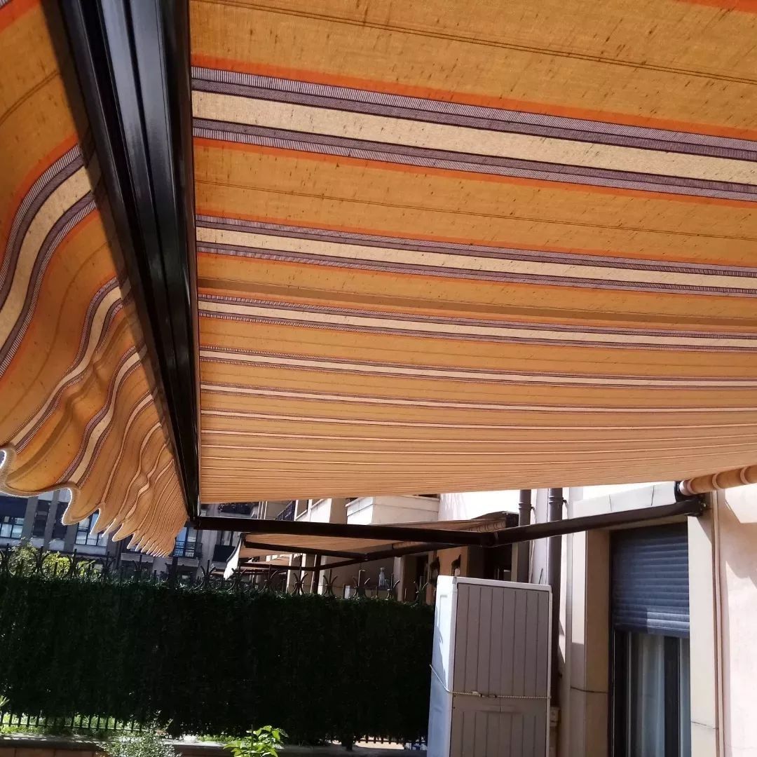 Un toldo a rayas, en tonos naranja, amarillo y marrón, se extiende sobre un espacio exterior, posiblemente un patio, proyectando sombra.