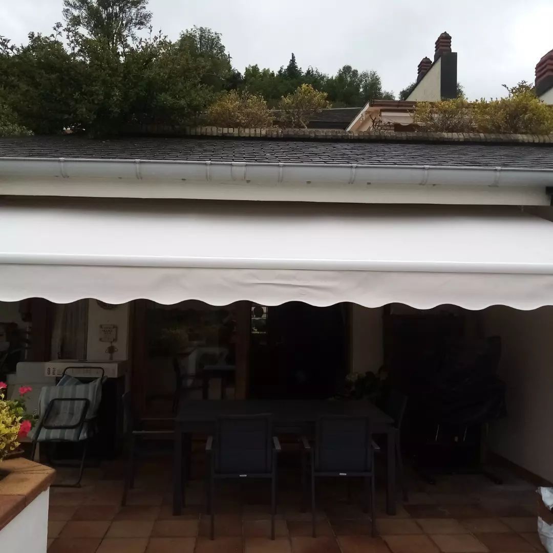 Un toldo blanco da sombra a un patio con mesa y sillas. Al fondo se ven un techo de tejas y árboles.
