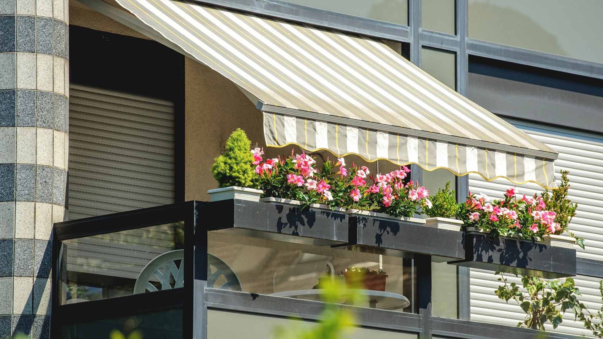 Balcón con toldo de rayas, jardineras con flores rosas y vista a la fachada de un edificio.