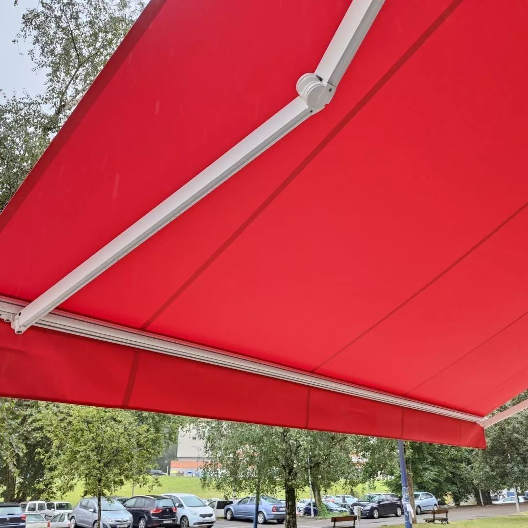 Toldo retráctil rojo extendido, que da sombra a un estacionamiento. Marco blanco, árboles y autos visibles.