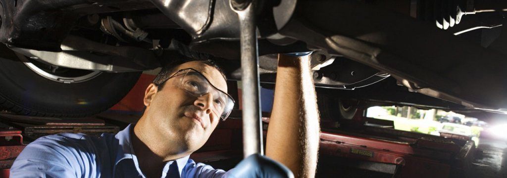 Mecánico trabajando debajo de un coche, usando una llave inglesa. Lleva gafas y guantes de seguridad.