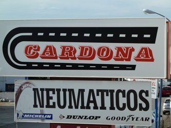 Letrero para “Cardona” (texto en rojo) con gráfica de carretera, y “Neumaticos” (texto en negro) con logotipos de marcas de neumáticos.