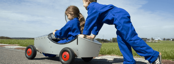 Dos niños con monos azules, uno empuja un pequeño coche gris mientras el otro va sentado en él sobre una pista pavimentada.