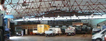 Gran interior de almacén con camiones estacionados, vigas metálicas expuestas y luces en el techo.