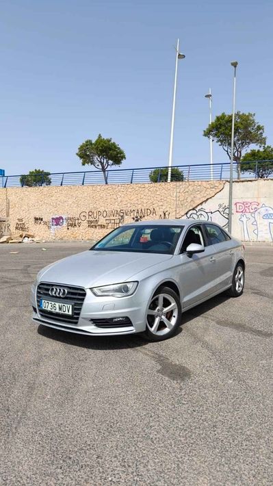Un sedán plateado estacionado en un terreno baldío junto a un muro cubierto de grafitis bajo un cielo azul despejado.