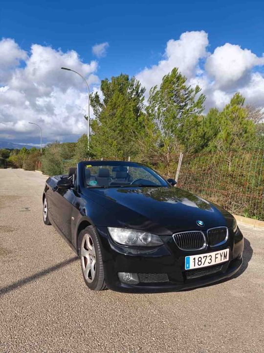 Un BMW negro aparcado en un camino de grava, con árboles y un cielo azul nublado al fondo.