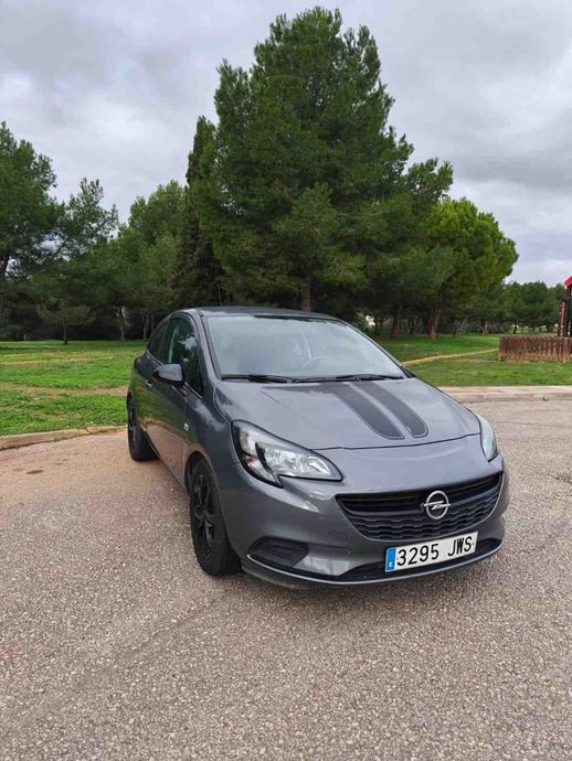 Un hatchback gris estacionado en un terreno de grava con árboles y césped al fondo.
