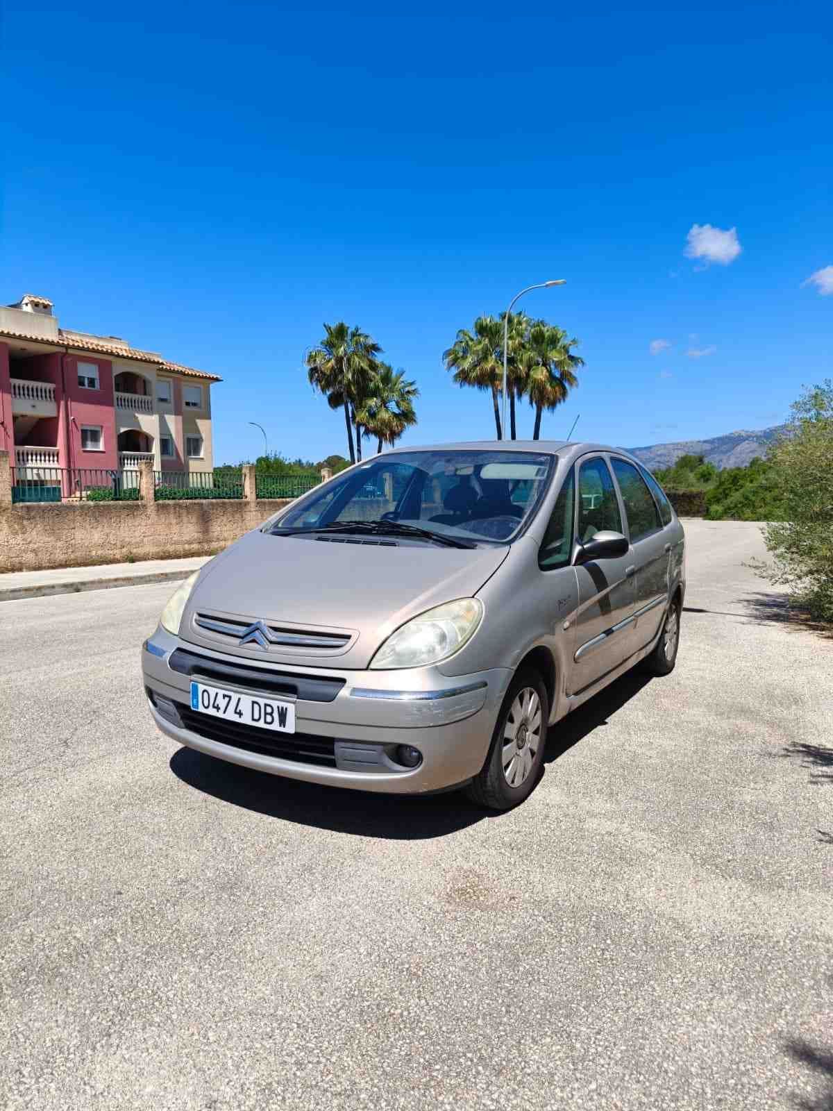 Un hatchback plateado estacionado en una carretera soleada junto a palmeras y edificios bajo un cielo azul.