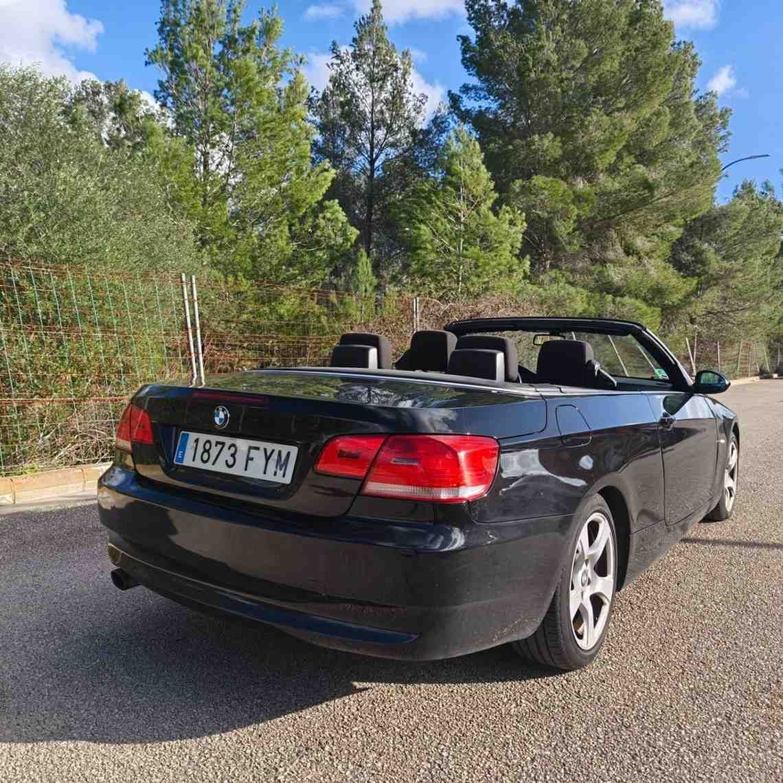 Coche descapotable negro aparcado al borde de la carretera con árboles al fondo, vista trasera.