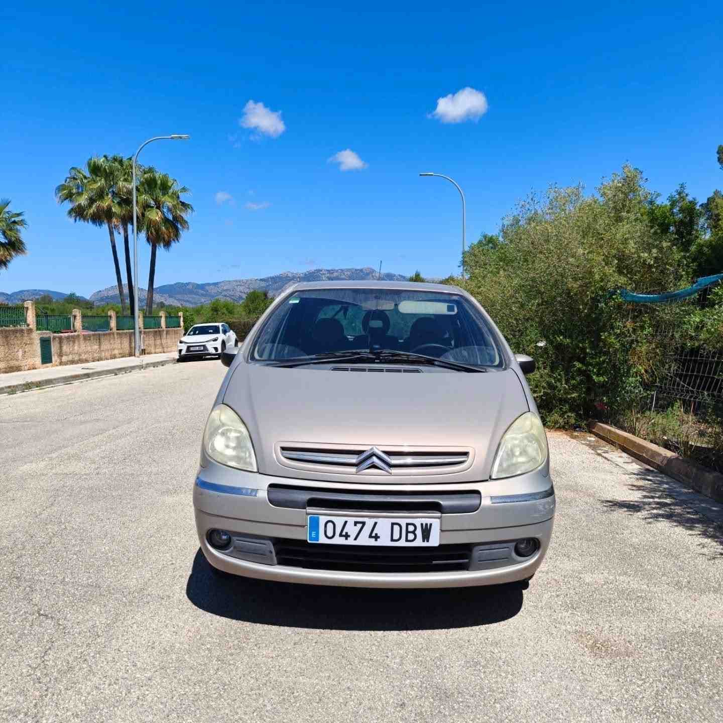 Un coche plateado aparcado en una carretera soleada con palmeras y cielo azul de fondo.