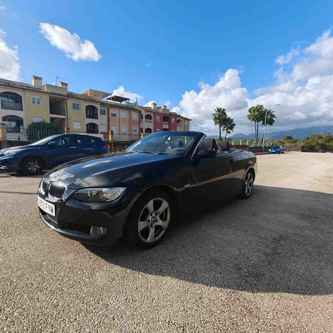 Coche deportivo descapotable negro aparcado en un soleado terreno de grava cerca de apartamentos y montañas bajo un cielo azul.