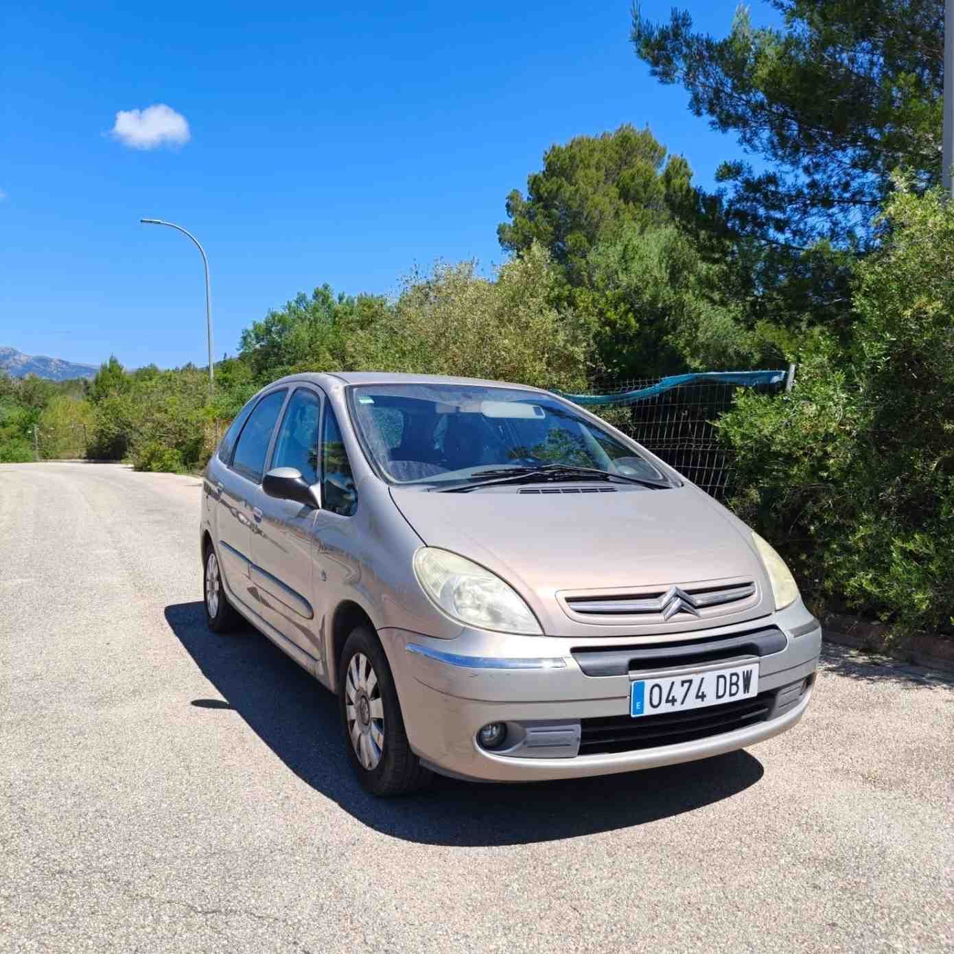 Un coche compacto de color beige aparcado en una carretera soleada, con árboles y cielo azul de fondo.