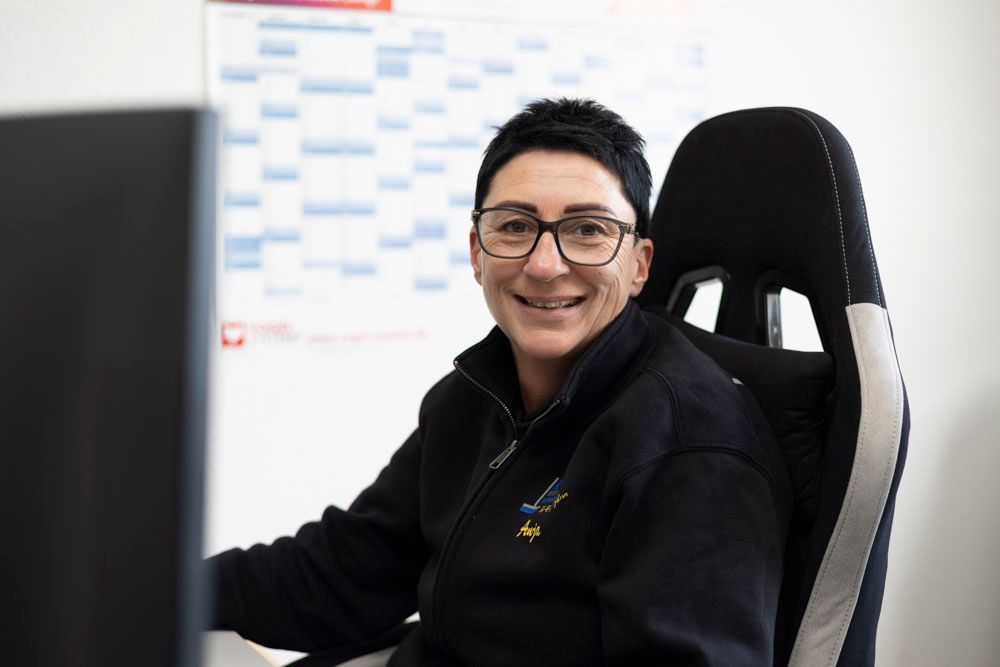 Eine Person lächelt an einem Schreibtisch in einem Büro; sie trägt eine Brille und eine schwarze Jacke; im Hintergrund ist eine Tafel zu sehen.