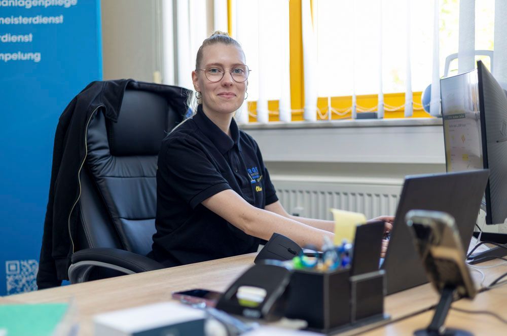 Eine Person sitzt an einem Schreibtisch in einem Büro und lächelt in die Kamera; Laptop und Monitor sind im Bild.