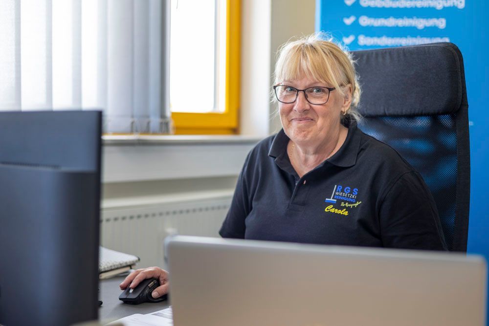 Eine Frau sitzt mit einem Laptop an einem Schreibtisch in einem hellen Büro und lächelt in die Kamera.