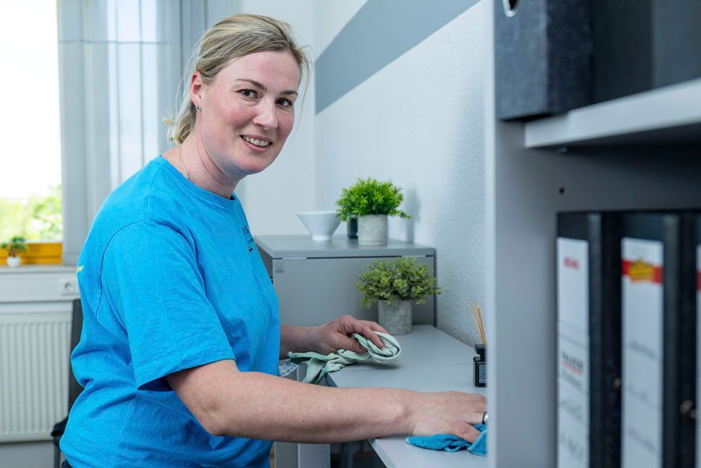 Eine Frau in einem blauen Hemd arbeitet an einem Schreibtisch in einem Büro, benutzt eine Computermaus und schaut in die Kamera.