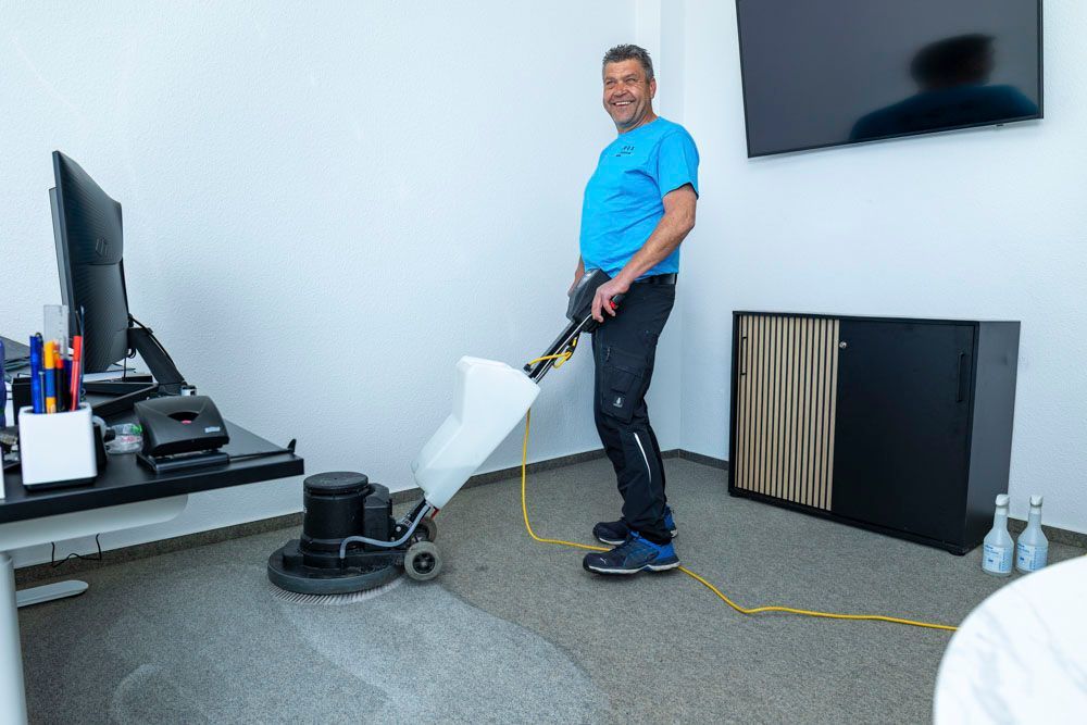 Ein Mann saugt den Büroboden neben einem Schreibtisch und einem an der Wand montierten Fernseher.