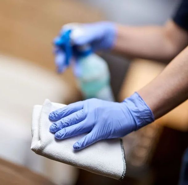 Hände in blauen Handschuhen, die eine Oberfläche besprühen und mit einem Tuch abwischen.