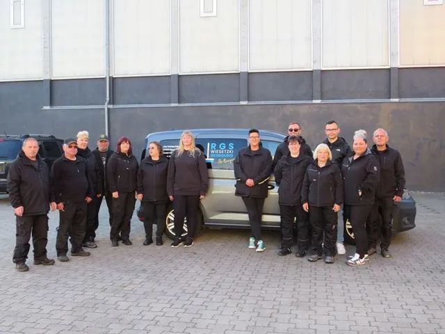 Eine Gruppe von Personen in schwarzen Uniformen posiert vor einem Lieferwagen mit Text. Im Hintergrund ist ein Gebäude zu sehen.